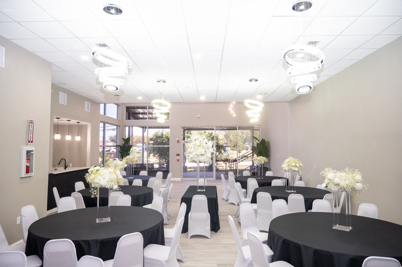 Decorated Onyx Event Center main hall with black table linens, white chairs, tall white floral centerpieces, ring chandeliers, and front windows